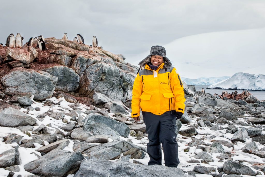 black woman in yellow Quark Expedition jacket standing on rocks with a group of dirty penguins behind her
