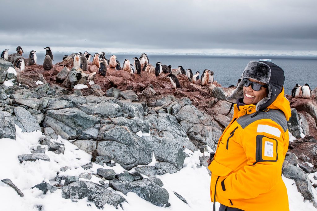 woman smiling next to a large group of penguins with poop all over the rocks beneath them