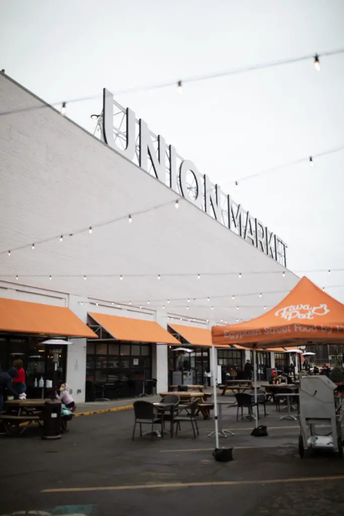 A photo of the front of the Union Market on a gloomy cloud-covered day. The Union Market sign at the top of the building. At the bottom of the image are tables and chairs where patrons can sit and eat their meals. 