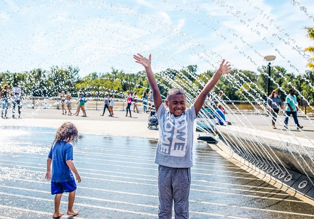 Young boy standing under sprinkler with arms stretched upward as water sprays and kids play around him.