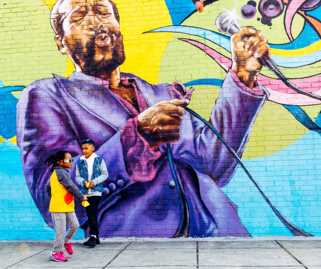 Two kids playing in front of a large colorful mural of Marvin Gaye singing. 