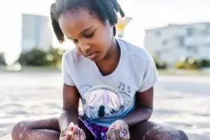 close up of little girl sitting on beach planing with sea shells and sand.