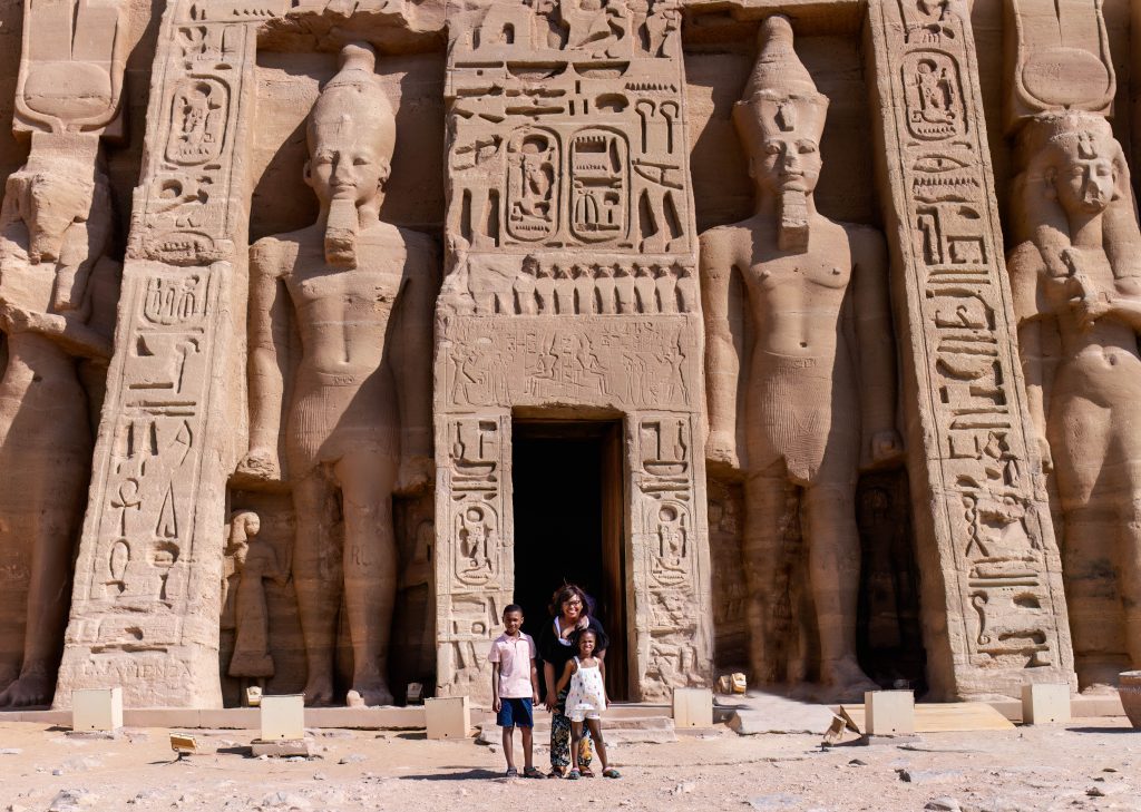 Woman with two children standing in front of large Egyptian engraving in the side of a mountain.