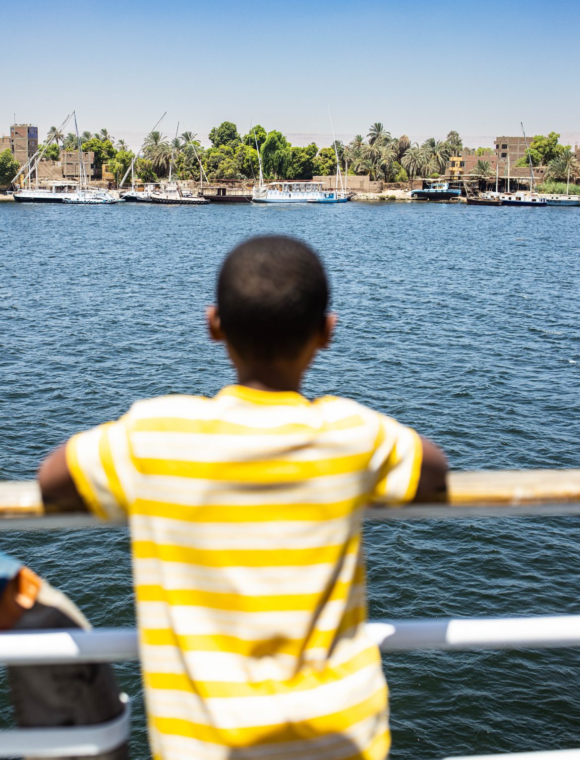 Little boy in foreground looking out at the Nile River and banks of the river from the cruise boat.